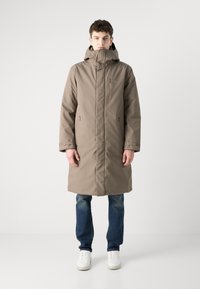 Lang, beige kåpe med høy krage og justerbar hette. Har glidelåslommer, solid tekstur og rett snitt. Brukes med blå jeans.