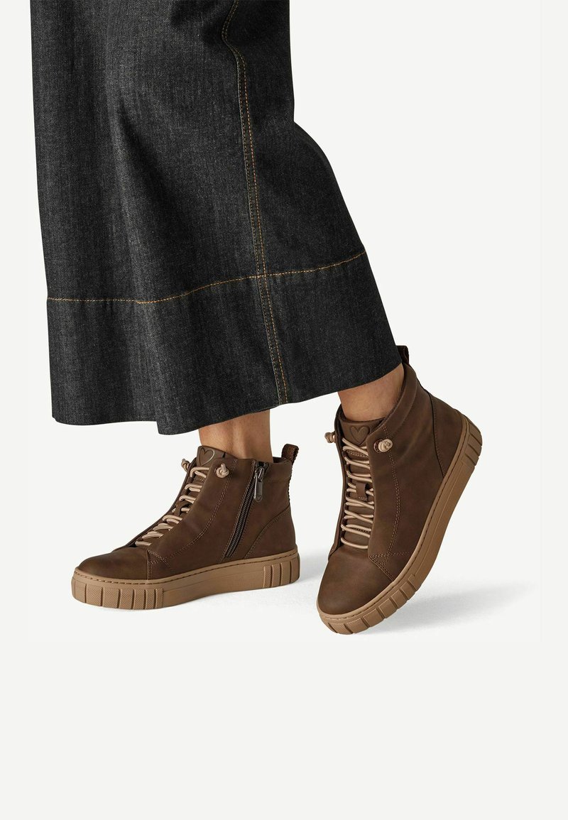 Personne portant un jean large foncé et des baskets montantes marron avec fermeture éclair latérale et lacets beige, un pied légèrement levé.
