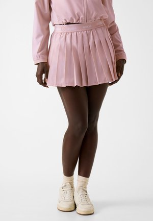Kvinde iført en rosa plisseret nederdel med GUESS-logo i taljen, matchende rosa jakke, cremefarvede sokker og hvide GUESS-sneakers, stående med benene krydsede.
