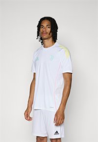 Fehér sportmez rövid ujjakkal és rózsaszín díszítéssel. Sárga Adidas csíkokkal a vállakon. Légáteresztő anyagból készült.