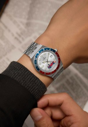 Silver watch with a round white dial, blue and red bezel, and patterned automatic movement. Attached to a stainless steel bracelet.