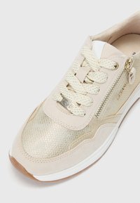 Baskets beiges avec un upper métallique doré texturé, des lacets crème, une fermeture éclair sur le côté et une semelle en caoutchouc blanche à bout rond.