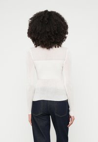 Bela rebrasta majica s dolgimi rokavi in oprijetim krojem. Prosojna tekstura. V kombinaciji s temno modrimi kavbojkami. Ni vidnih dodatnih podrobnosti.