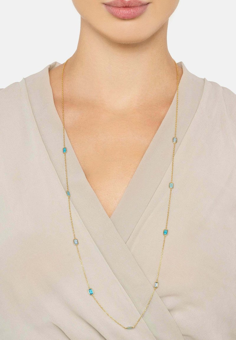 Gold chain necklace featuring turquoise rectangular stones evenly spaced along the length, set against a light-colored fabric backdrop.