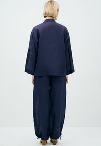 Completo in lino blu navy composto da due pezzi, caratterizzato da una tunica ampia con maniche larghe e pantaloni abbinati con cordoncino in vita. Texture liscia in tutto il completo.