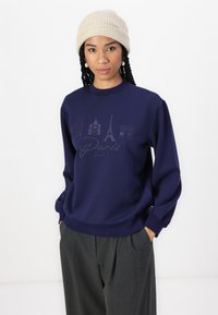 Sweatshirt azul marinho com marcos de Paris impressos em cinza escuro, apresenta um decote redondo e mangas longas. Combinado com calças cinzas.