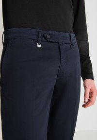 Pantaloni blu navy realizzati in tessuto morbido, con chiusura a bottone, passanti per cintura e una leggera lucentezza. Dettaglio di cucitura visibile sulle tasche.