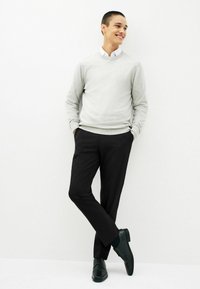 Lys grå sweater med rund hals, sorte bukser og sorte lædersko. Personen står med hænderne i lommerne mod en hvid baggrund.