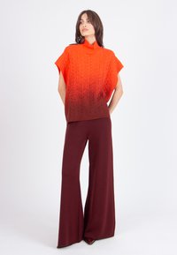 Woman wearing sleeveless orange to brown ombre turtleneck sweater and wide-leg burgundy pants, standing against a plain white background.