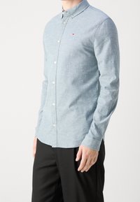 Mies, joka on pukeutunut vaaleansiniseen napitettyyn pitkähihaiseen paitaan, jossa on pieni logo rinnassa, ja mustiin housuihin, seisoo valkoista taustaa vasten.