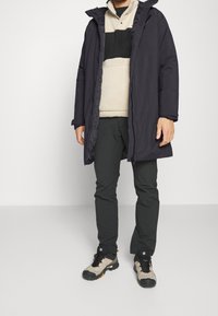 Mörkblå ytterjacka med huva, i kombination med en beige och svart färgblockerad fleecepullover och svarta byxor. Ljusbruna och svarta vandringsskor.