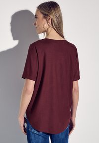 Burgundy kortärmad topp med uppvikta ärmar, mjukt tyg, rundad nederkant, bärs med ljusblå jeans, visad bakifrån.