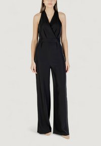 Donna che indossa un jumpsuit senza maniche nero con collo all'americana e gambe larghe, con scarpe a punta nuda, in piedi su uno sfondo neutro.