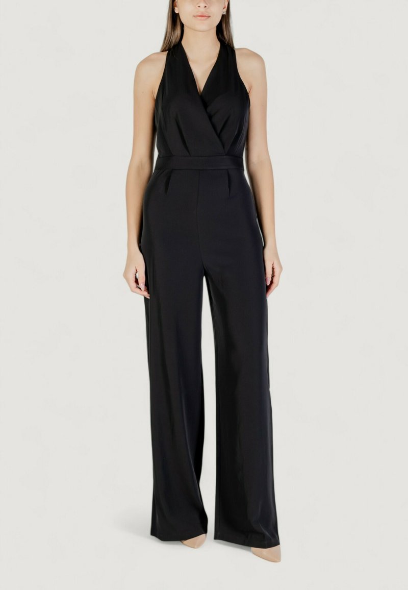 Donna che indossa un jumpsuit senza maniche nero con collo all'americana e gambe larghe, con scarpe a punta nuda, in piedi su uno sfondo neutro.