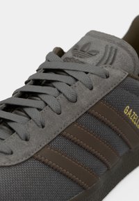 Γκρι παπούτσια Adidas Gazelle με υφασμάτινο επάνω μέρος, καφέ δερμάτινες ρίγες και γκρι κορδόνια. Διαθέτουν καουτσούκ σόλα και ανάγλυφο λογότυπο στη γλώσσα.
