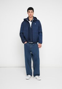 Marineblauer Steppmantel mit horizontalen Nähten, getragen über einem marineblauen Hoodie und einem weißen Polohemd. Kombiniert mit locker sitzenden blauen Jeans und weißen Turnschuhen.