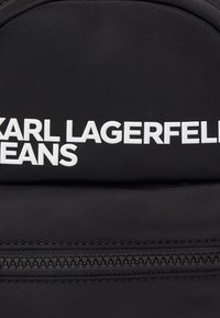 Black fabric with white raised "KARL LAGERFELD JEANS" logo above a black zipper on a bag or backpack.