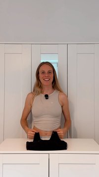 Lys grå ermeløs topp med rund hals, kombinert med et svart tekstilobjekt på en hvit overflate. Bakgrunnen har enkel hvit skapinnredning.