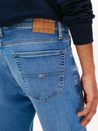 Svijetloplave traperice s kožnom oznakom na kojoj piše "TOMMY JEANS." Stražnji džep ima brendirani detalj s crveno-bijelo-plavim logotipom.