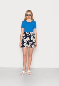 Camisa azul curta, saia floral azul-marinho, sandálias brancas; modelo a usar óculos de sol, em pé sobre um chão cinza claro contra um fundo simples.
