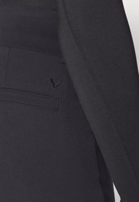 Svart skreddersydd jakke med glatt stoff, strukturert design og en subtil brodert logo detalj på lommen.