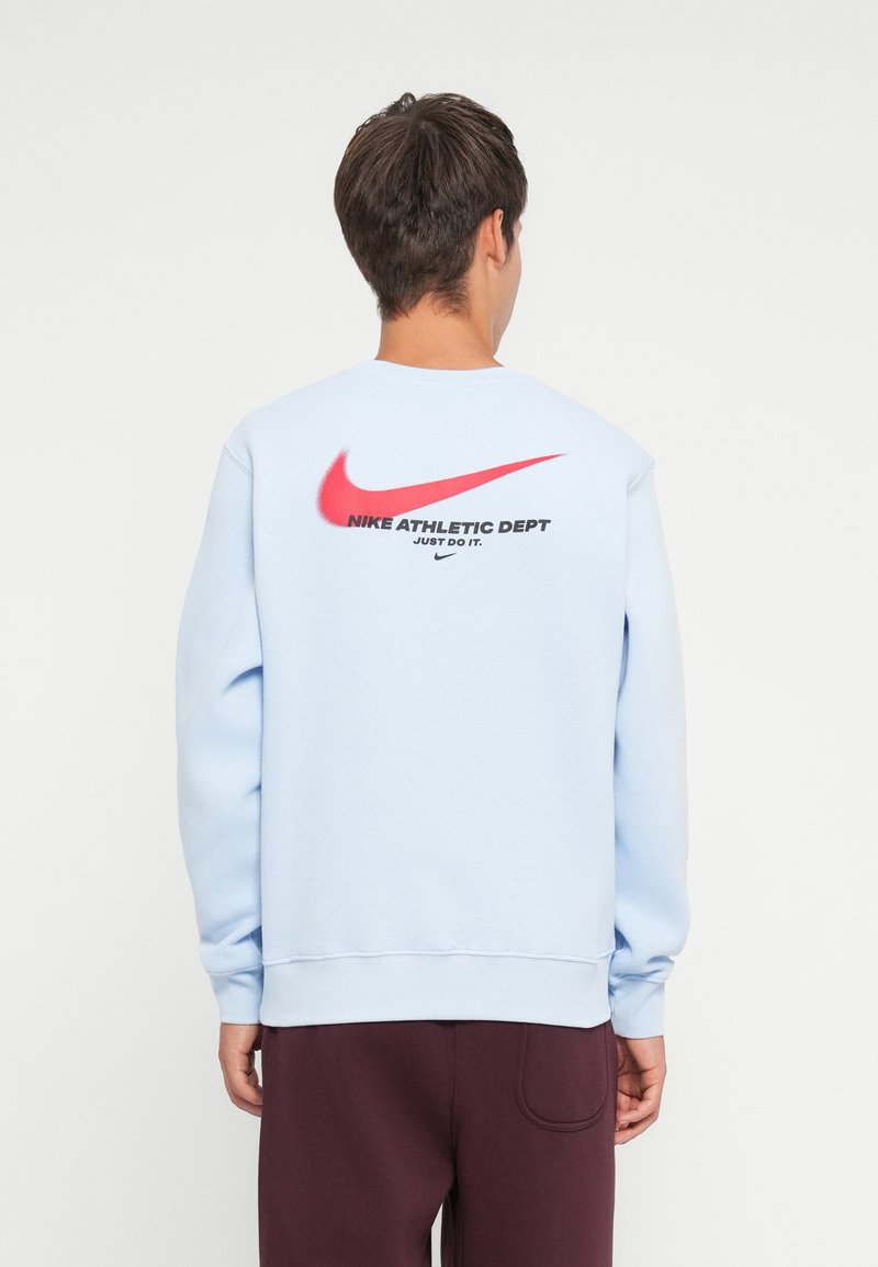 Lichtblauwe sweatshirt met een ronde hals, voorzien van een grote rode Nike Swoosh en de tekst "Nike Athletic Dept. Just Do It." op de achterkant.