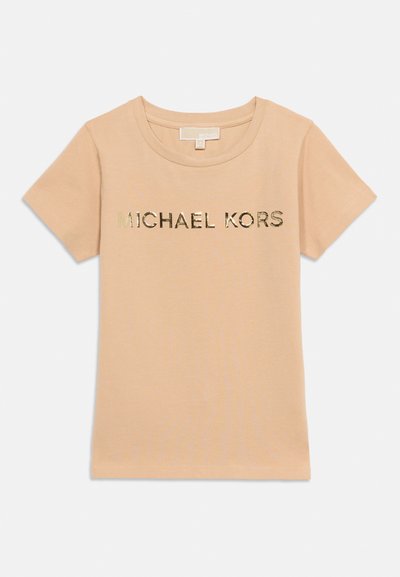 Béžové tričko s krátkym rukávom a zlatým kovovým logom "MICHAEL KORS" umiestneným v strede na hrudi, okrúhly výstrih, jednoduché pozadie.