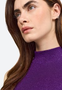 Haut sans manches violet avec un col montant, en tissu texturé et scintillant. Longs cheveux bruns partiellement visibles sur un fond clair.