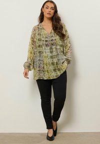 Fließende grün-lila gemusterte Bluse mit tiefem V-Ausschnitt, langen Ärmeln und plissierten Details, kombiniert mit schwarzen Skinny-Jeans und schwarzen Ballerinas.