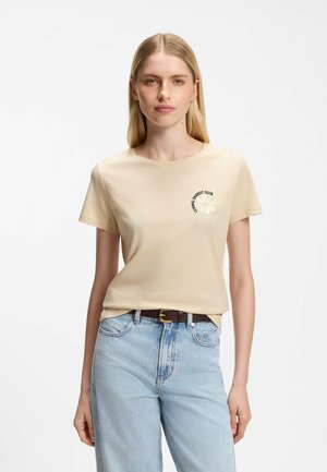 Kobieta z prostymi blond włosami ma na sobie beżowy t-shirt z napisem „Cosmic Energy Club” i grafiką stokrotki, włożony w jasnoniebieskie dżinsy.