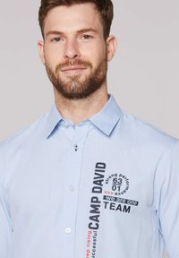 Hellblaues Hemd mit Knopfverschluss aus strukturiertem Stoff, mit Punktkragen und bedruckten Akzenten in Schwarz und Rot mit dem Text "CAMP DAVID TEAM."