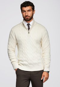 Mężczyzna z brodą, ubrany w biały sweter z dzianiną w splot warkoczowy z pół zamkiem, na białej koszuli i w paskowaną krawat, ciemne spodnie oraz zegarek na lewym nadgarstku.