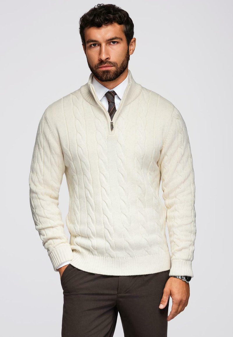 Mężczyzna z brodą, ubrany w biały sweter z dzianiną w splot warkoczowy z pół zamkiem, na białej koszuli i w paskowaną krawat, ciemne spodnie oraz zegarek na lewym nadgarstku.