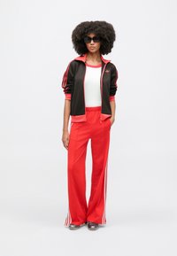 Czerwony dres z czarną kurtką na zamek z czerwonymi akcentami i białymi paskami. Pod spodem biały top. Modelka nosi ciemne okulary przeciwsłoneczne.