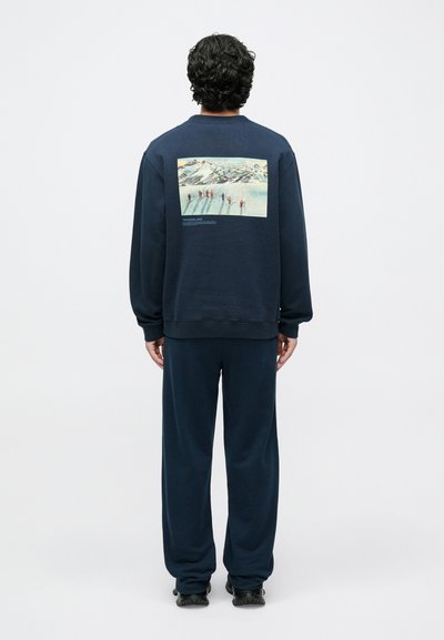 Temno modra majica z dolgimi rokavi in okroglim izrezom, na hrbtni strani s pravokotnim grafičnim potiskom, ki prikazuje zasneženo pokrajino z osebami.