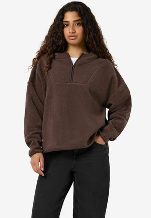 Kvinde med langt krøllet hår iført en brun fleece-pullover med kvart lynlås og sorte bukser, stående med den ene hånd i lommen.