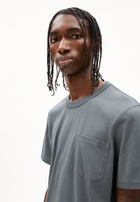 Grijze katoenen t-shirt met een relaxte pasvorm, ronde hals en een borstzak. De stof heeft een gladde textuur, en de stiksels zijn zichtbaar op de randen.