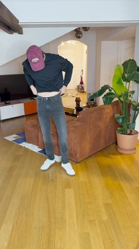 Svart hettegenser, grå slim-fit jeans og hvite sneakers. Rom med tregulv, en brun sofa, en plante og en TV i bakgrunnen.