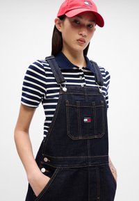 Tumši džinsi ar regulējamām siksnām un priekšējo kabatu. Apvienoti ar tumši zilu un baltu svītrotu polo krekls un sarkanu cepuri.