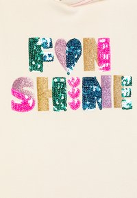 Un tissu couleur crème arbore le texte "FUN SHINE" en sequins multicolores et vibrants. Les détails incluent des cœurs et différentes textures dans les lettres.