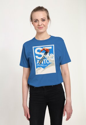 Młoda kobieta w niebieskiej koszulce z grafiką narciarza skaczącego na zaśnieżonym stoku oraz napisem "St. Anton", stojąca na jednolitym tle.