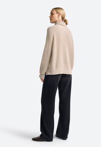 Beige gestrickter Pullover mit hohem Kragen, leicht übergroßer Passform und gerippter Textur, kombiniert mit weiten schwarzen Hosen und flachen Schuhen.