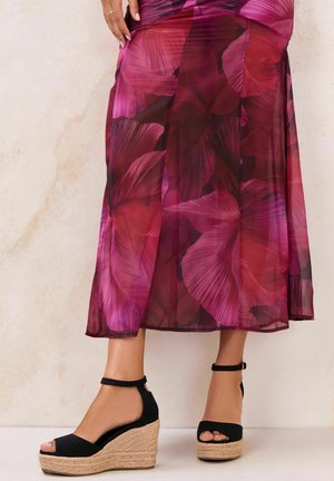 Lower legs wearing black ankle-strap wedge sandals paired with a sheer floral midi skirt in shades of red and purple.