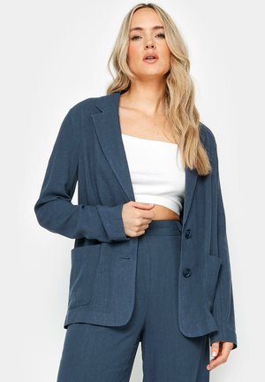 Blazer blu scuro con taglio relaxed, due tasche frontali e due bottoni. Indossato sopra un top corto bianco, caratterizzato da un tessuto testurizzato.