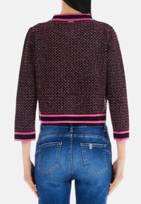 Giacca cropped nera e rosa con un motivo testurizzato, caratterizzata da una larga striscia rosa lungo l'orlo e le maniche. Indossata sopra jeans blu.