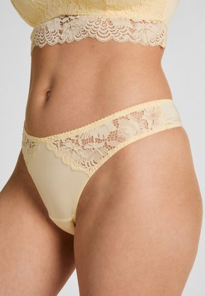 Frau trägt hellgelbe Spitzendessous mit Blumenmuster, fokussiert auf Mittelteil und Hüftbereich vor einem schlichten hellen Hintergrund.