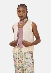 Chaleco de encaje en color crema claro con bajo de flecos, superpuesto a una blusa lavanda y pantalones de estampado floral con varios colores y texturas.