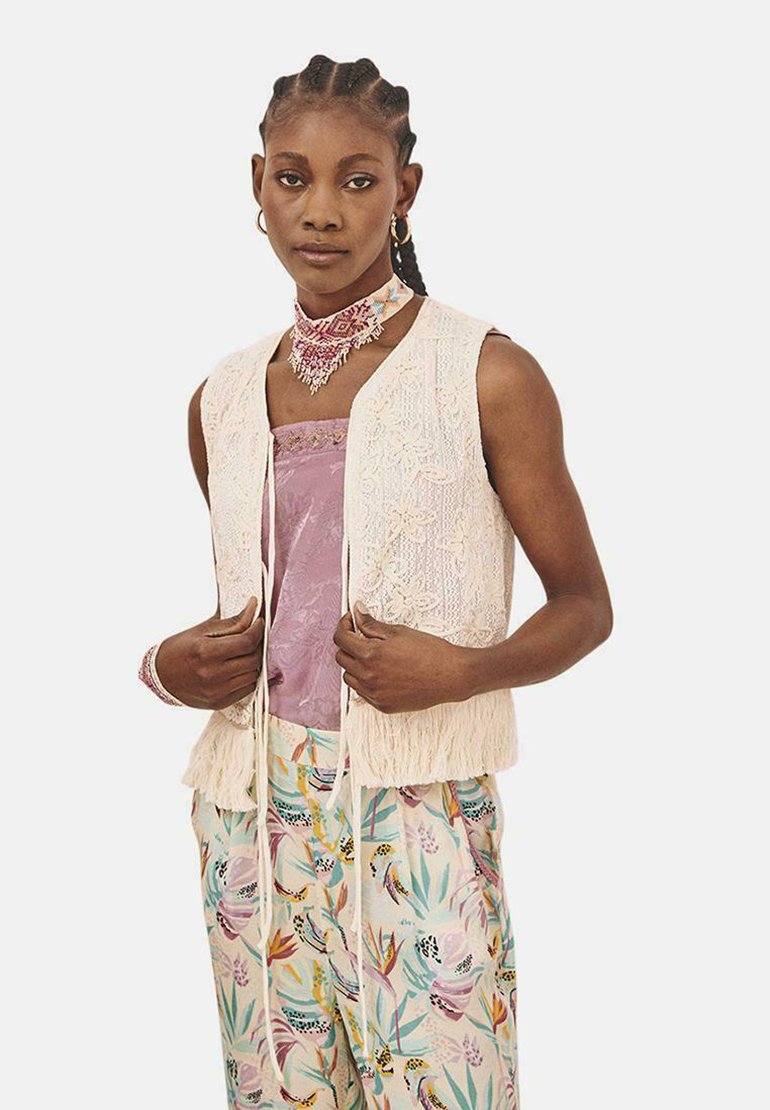 Chaleco de encaje en color crema claro con bajo de flecos, superpuesto a una blusa lavanda y pantalones de estampado floral con varios colores y texturas.
