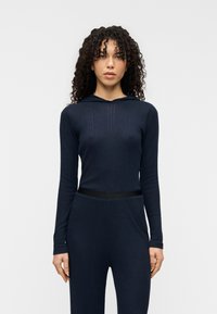 Μπλε σκούρο μακρυμάνικο φούτερ με ριμπ υφή και λεπτομέρειες πλευρικής επένδυσης. Διαθέτει μαύρη ελαστική ζώνη στη μέση, εφαρμοστή σιλουέτα και κουκούλα.