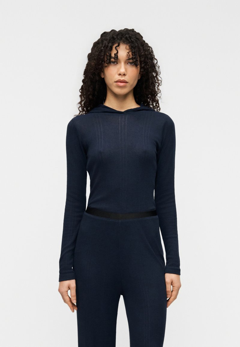 Μπλε σκούρο μακρυμάνικο φούτερ με ριμπ υφή και λεπτομέρειες πλευρικής επένδυσης. Διαθέτει μαύρη ελαστική ζώνη στη μέση, εφαρμοστή σιλουέτα και κουκούλα.